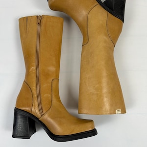 Puede incluir: Un par de botas de cuero color canela con tacones cuadrados negros. Una bota est&aacute; de pie, la otra est&aacute; en &aacute;ngulo. Ambas botas tienen punta cuadrada y cremallera en el interior. Las botas tienen una peque&ntilde;a etiqueta en el lateral.