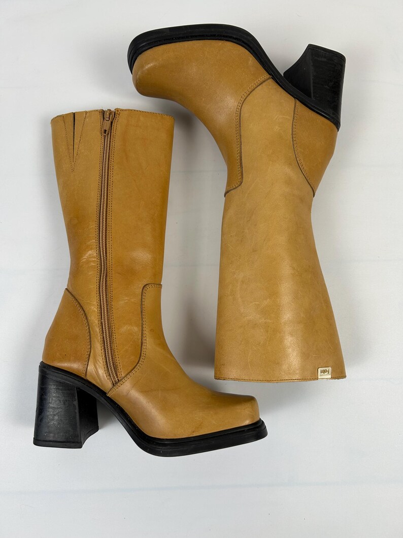 Puede incluir: Un par de botas de cuero color canela con tacones cuadrados negros. Una bota est&aacute; de pie, la otra est&aacute; en &aacute;ngulo. Ambas botas tienen punta cuadrada y cremallera en el interior. Las botas tienen una peque&ntilde;a etiqueta en el lateral.