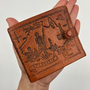 May include: A brown leather wallet with a map of Australia embossed on the front. The wallet features a snap closure and the word "AUSTRALIA" in a banner. The map includes illustrations of landmarks and cities.