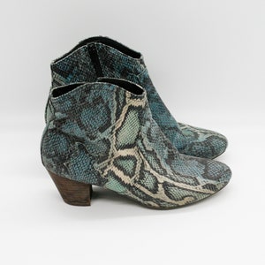 Könnte beinhalten: Knöchelstiefel mit Schlangenmuster in Türkis, Blau und Creme. Die Stiefel haben eine spitze Zehenpartie, einen niedrigen Holzabsatz und einen Reißverschluss an der Seite. Die Stiefel stehen vor einem weißen Hintergrund. Absatzhöhe ca. 5 cm.
