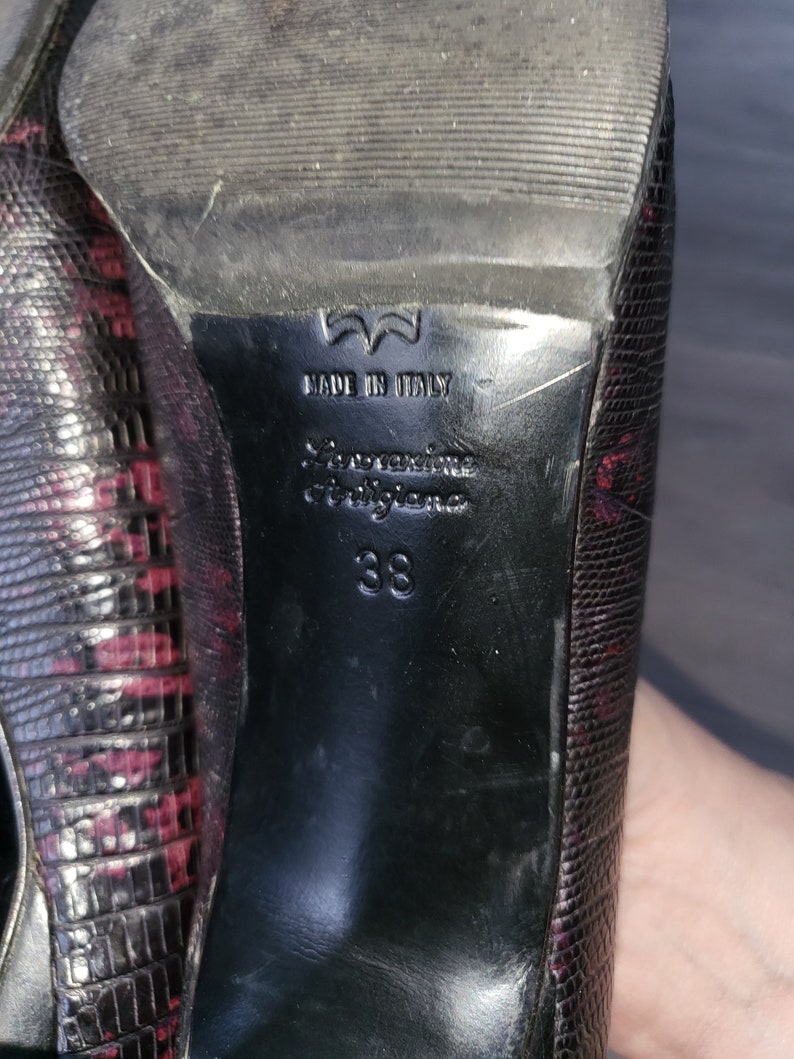 May include: Black leather heels with a snakeskin pattern and a red undertone. The heels have a label that reads "Made in Italy" and "Salvatore Ferragamo" with the size "38" printed below.