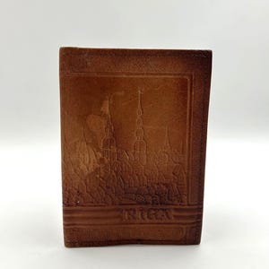 May include: A brown leather wallet with an embossed cityscape design. The wallet features a detailed depiction of buildings and spires, with the city name "RIGA" at the bottom. The wallet is rectangular and appears to be closed.