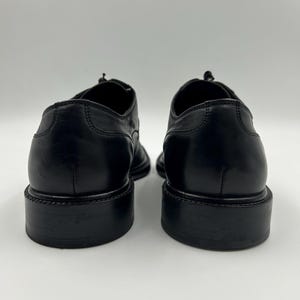 K&ouml;nnte beinhalten: Ein Paar schwarze Lederschuhe, von hinten gesehen. Die Schuhe haben ein klassisches Design mit runder Spitze und einem gestapelten Absatz. Das Leder wirkt glatt und poliert, mit sichtbaren Nahtdetails.