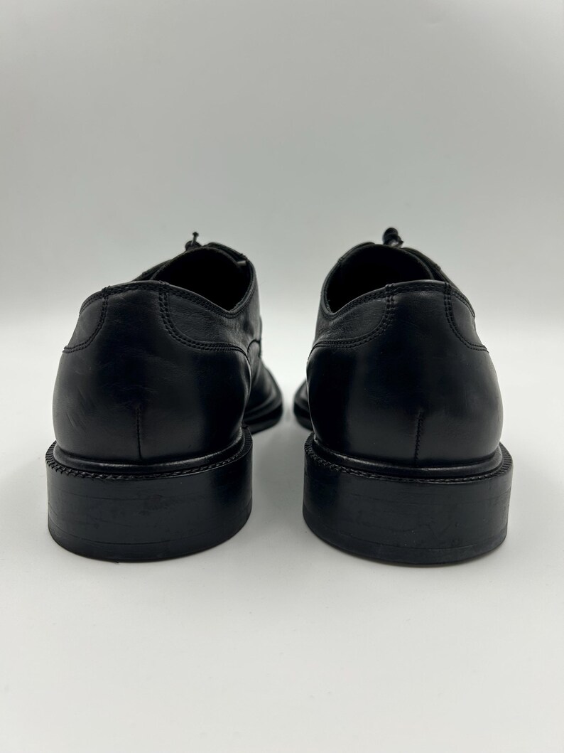 K&ouml;nnte beinhalten: Ein Paar schwarze Lederschuhe, von hinten gesehen. Die Schuhe haben ein klassisches Design mit runder Spitze und einem gestapelten Absatz. Das Leder wirkt glatt und poliert, mit sichtbaren Nahtdetails.