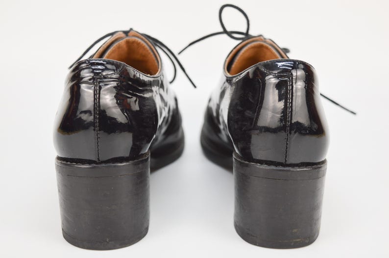 Puede incluir: Par de zapatos de tac&oacute;n de charol negro con cierre de cordones. Los zapatos tienen un tac&oacute;n grueso y una puntera redonda.