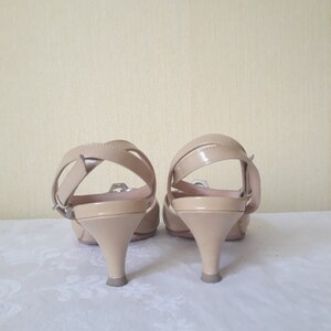 May include: A pair of beige heeled sandals with a strap across the ankle and a buckle closure. The sandals have a low heel and are made of a smooth, shiny material.