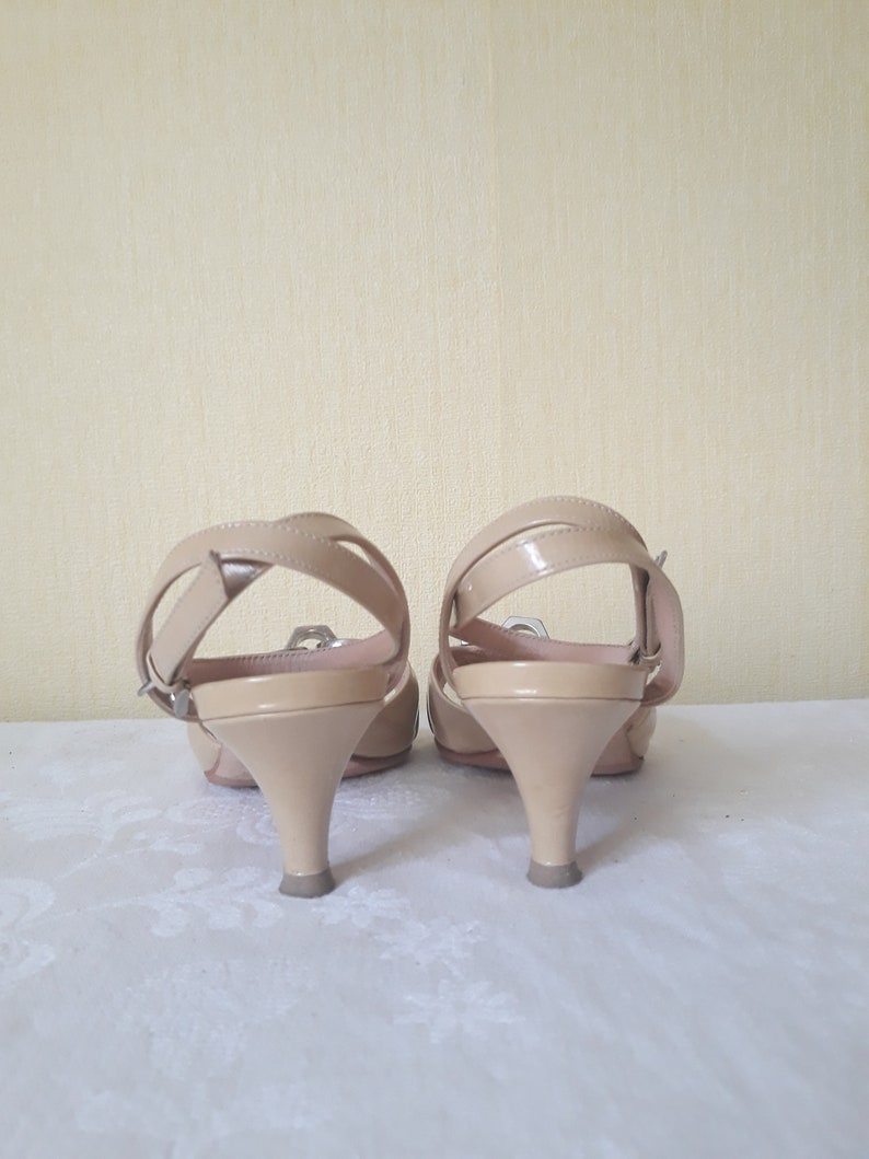 May include: A pair of beige heeled sandals with a strap across the ankle and a buckle closure. The sandals have a low heel and are made of a smooth, shiny material.