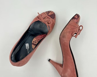 May include: A pair of pink suede high heels with a snakeskin pattern on the toe. The heels are about 3 inches tall and have a bow on the front.