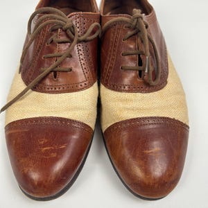 May include: Pair of two-tone oxford shoes. The shoes feature brown leather accents on the toe and heel, with a tan fabric midsection. Brown laces and a classic design complete the look. The shoes are displayed on a white background.