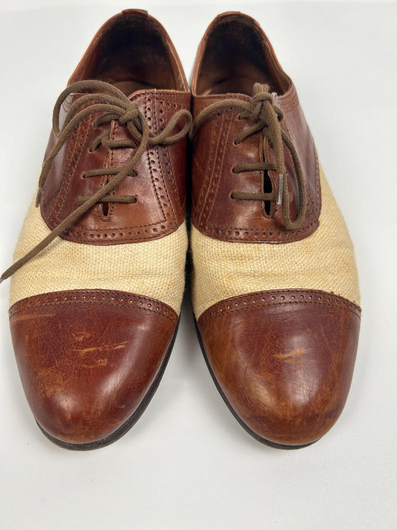 May include: Pair of two-tone oxford shoes. The shoes feature brown leather accents on the toe and heel, with a tan fabric midsection. Brown laces and a classic design complete the look. The shoes are displayed on a white background.
