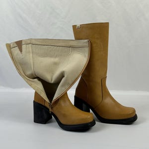 Puede incluir: Un par de botas de cuero color canela con tacones cuadrados negros. Una bota est&aacute; desabrochada, revelando un interior beige. Las botas tienen punta cuadrada y cierre de cremallera. Sobre fondo blanco.