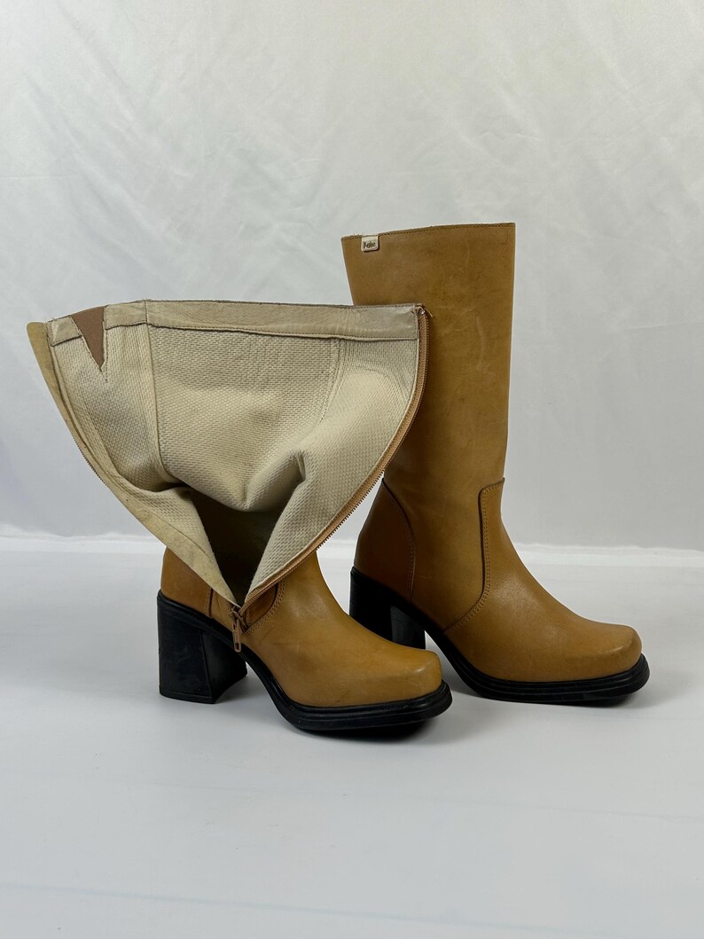 Puede incluir: Un par de botas de cuero color canela con tacones cuadrados negros. Una bota est&aacute; desabrochada, revelando un interior beige. Las botas tienen punta cuadrada y cierre de cremallera. Sobre fondo blanco.
