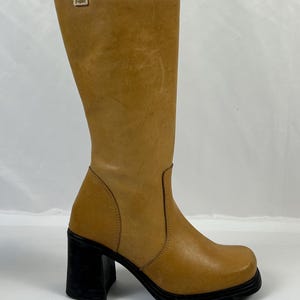 Puede incluir: Bota alta hasta la rodilla de cuero marr&oacute;n claro con puntera cuadrada y tac&oacute;n de bloque negro. La bota tiene un acabado liso, ligeramente texturizado y una peque&ntilde;a etiqueta con el logotipo en la parte superior. El tac&oacute;n mide unos 7.6 cm.