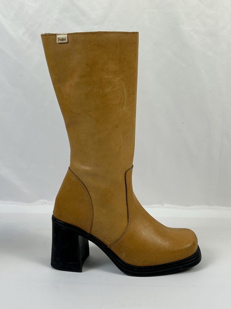 Puede incluir: Bota alta hasta la rodilla de cuero marr&oacute;n claro con puntera cuadrada y tac&oacute;n de bloque negro. La bota tiene un acabado liso, ligeramente texturizado y una peque&ntilde;a etiqueta con el logotipo en la parte superior. El tac&oacute;n mide unos 7.6 cm.