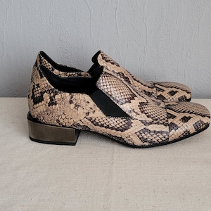 Puede incluir: Un par de zapatos de piel de serpiente marrón y negro con un panel elástico negro y un tacón grueso.