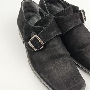 K&ouml;nnte beinhalten: Ein Paar schwarze Wildlederschuhe mit Schnalle. Die Schuhe haben eine quadratische Spitze und einen einzigen Schnallenriemen mit dem eingepr&auml;gten Wort "STILO" auf der Schnalle.