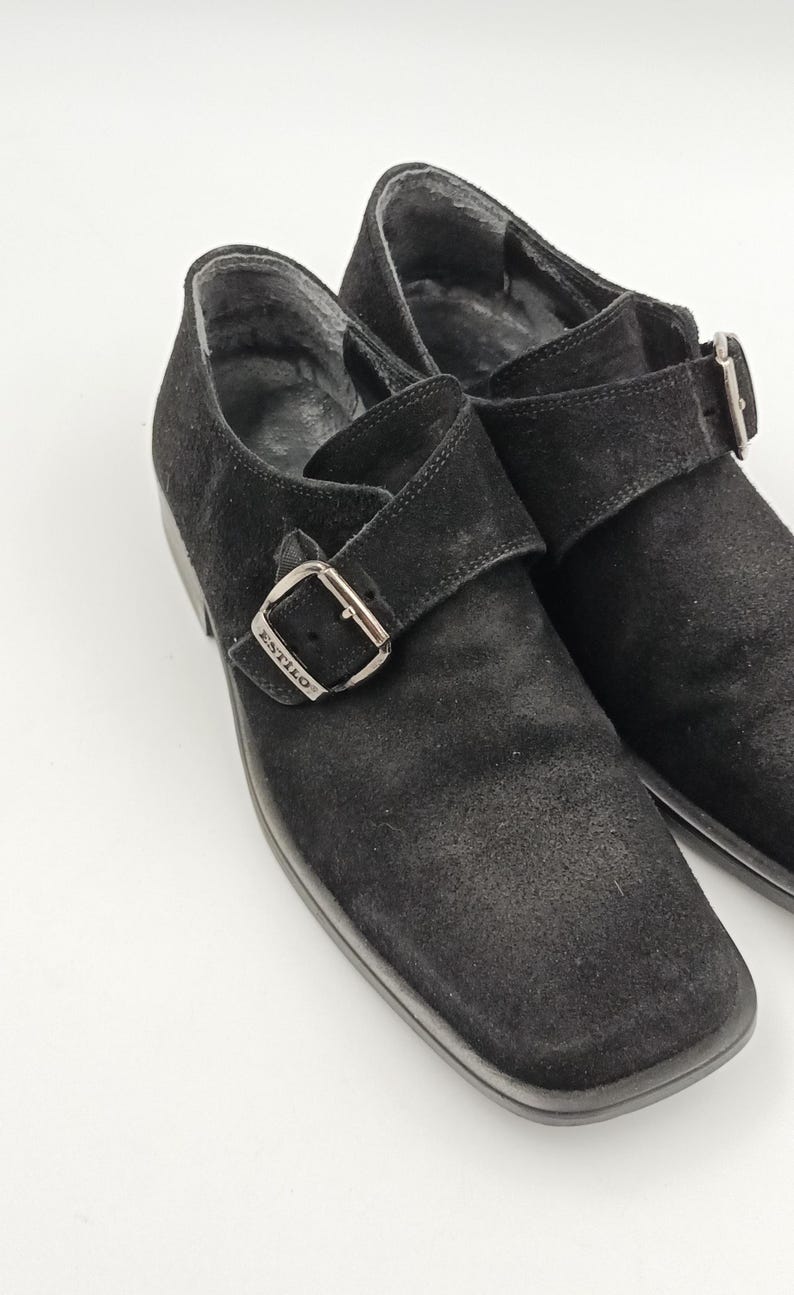 K&ouml;nnte beinhalten: Ein Paar schwarze Wildlederschuhe mit Schnalle. Die Schuhe haben eine quadratische Spitze und einen einzigen Schnallenriemen mit dem eingepr&auml;gten Wort "STILO" auf der Schnalle.