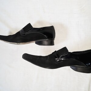 Puede incluir: Un par de zapatos de vestir de cuero negro con punta puntiaguda y detalle de hebilla. Un zapato es de ante y el otro es de charol.