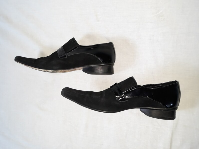 Puede incluir: Un par de zapatos de vestir de cuero negro con punta puntiaguda y detalle de hebilla. Un zapato es de ante y el otro es de charol.