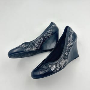 Può includere: Un paio di scarpe con zeppa nere con un rivestimento in pizzo floreale. I tacchi hanno una punta arrotondata e un piccolo dettaglio a forma di rosa sul davanti. Il tacco a zeppa è alto circa 7,6 cm.
