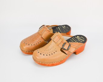 May include: A pair of tan leather clogs with a buckle strap. The clogs have a perforated design and orange soles. The clogs are made by the brand "Svend".