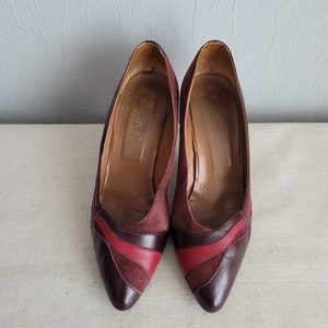 Puede incluir: Un par de zapatos de tacón de ante y cuero burdeos con detalles rojos. Los zapatos tienen punta puntiaguda y tacón bajo.