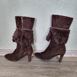May include: A pair of brown suede boots with a fur trim around the top of the boot. The boots have a high heel and a pointed toe.