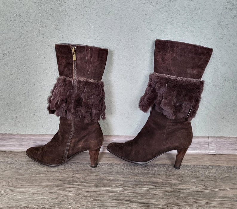 May include: A pair of brown suede boots with a fur trim around the top of the boot. The boots have a high heel and a pointed toe.