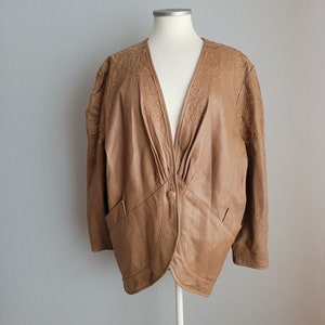 May include: A tan leather jacket with a V-neck design and a single button closure. The jacket features embroidered floral patterns on the shoulders and upper chest. It has two front pockets and is displayed on a mannequin.