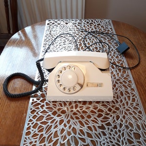 Può includere: Un telefono a disco bianco con un filo nero. Il telefono è appoggiato su un runner da tavolo argentato con un motivo floreale.