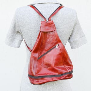 May include: A red leather backpack with a brown zipper and a brown strap. The backpack has a flap that closes with a buckle.