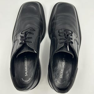 K&ouml;nnte beinhalten: Ein Paar schwarze Lederschuhe mit quadratischen Zehen und Schn&uuml;rverschluss. Der Markenname "SALAMANDER" ist auf der Innensohle aufgedruckt. Die Schuhe werden auf wei&szlig;em Hintergrund pr&auml;sentiert.