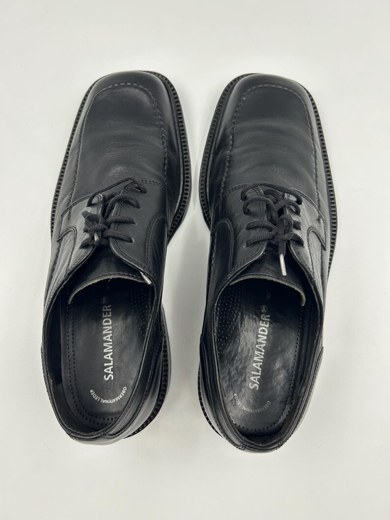K&ouml;nnte beinhalten: Ein Paar schwarze Lederschuhe mit quadratischen Zehen und Schn&uuml;rverschluss. Der Markenname "SALAMANDER" ist auf der Innensohle aufgedruckt. Die Schuhe werden auf wei&szlig;em Hintergrund pr&auml;sentiert.