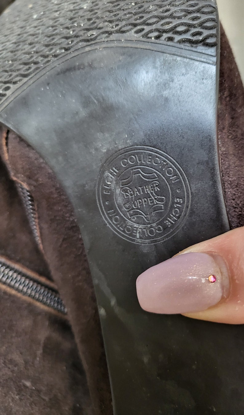 May include: Close-up of the brown suede heel of a boot with a black leather sole. The sole has a circular logo that reads "Elche Collection" and "Leather Upper".