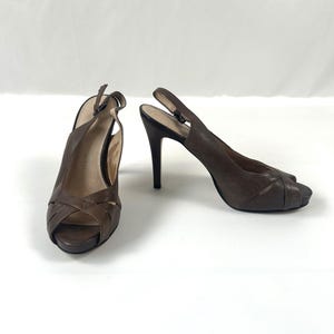Puede incluir: Un par de tacones slingback de cuero marrón. Los zapatos de punta abierta tienen un diseño entrecruzado sobre el pie y una fina correa que se abrocha alrededor del tobillo. Los tacones son de color marrón oscuro.