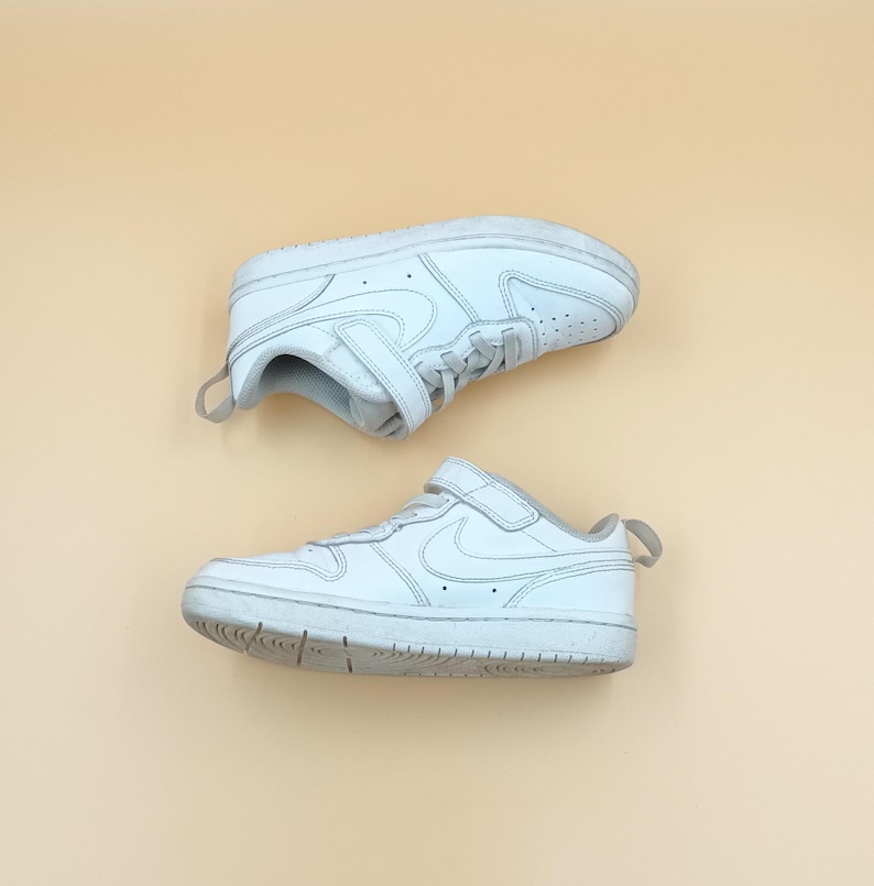 以下が含まれることがあります： ベルクロ留めの白い革のスニーカーのペア。スニーカーには側面に白いスウッシュロゴがあります。