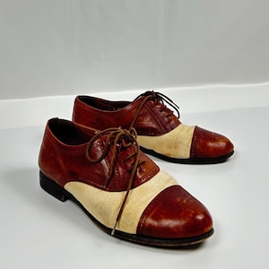 Può includere: Un paio di scarpe Oxford in pelle bicolore. Le scarpe sono marroni con una fascia color crema sul collo del piede. Le scarpe hanno lacci e una suola nera. Le scarpe sono esposte su una superficie bianca.