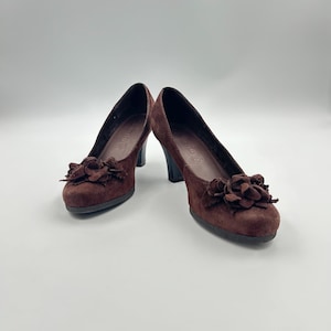 Könnte beinhalten: Ein Paar braune Wildlederschuhe mit Absatz und einer Blumenverzierung an der Spitze.