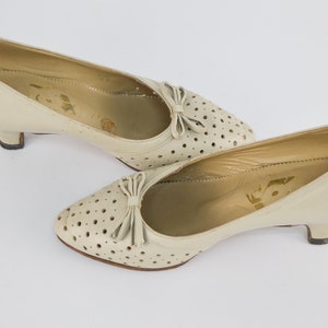 May include: A pair of off-white leather pumps with a small heel. The shoes have a perforated design and a bow detail on the toe.