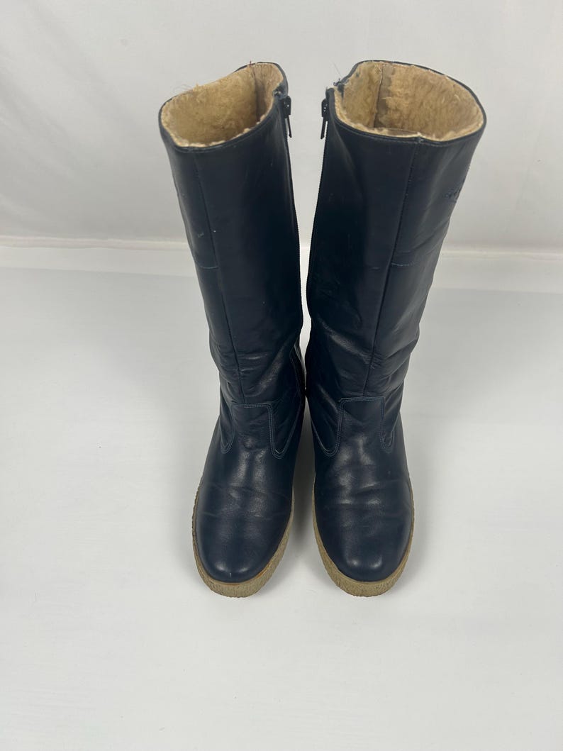 May include: A pair of navy blue knee-high boots with a beige faux fur lining. The boots have a zipper closure and a light brown sole. The boots are made of a smooth, possibly leather, material.