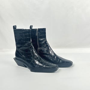 Könnte beinhalten: Ein Paar schwarze Stiefeletten mit einer Kunstleder-Krokodil-Textur. Die Stiefel haben eine quadratische Spitze, einen seitlichen Reißverschluss und einen niedrigen, abgewinkelten Absatz. Die Stiefel sind etwa 18 cm hoch.
