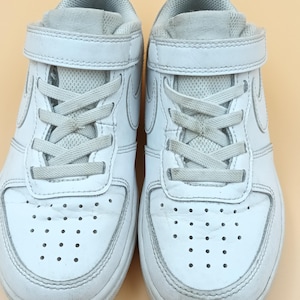 以下が含まれることがあります： ベルクロストラップとレースが付いた白い革のスニーカーのペア。スニーカーは白いソールと側面に白いスウッシュロゴが付いています。