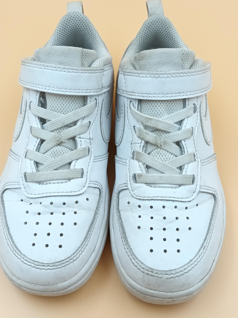 以下が含まれることがあります： ベルクロストラップとレースが付いた白い革のスニーカーのペア。スニーカーは白いソールと側面に白いスウッシュロゴが付いています。