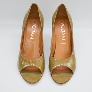 Puede incluir: Un par de tacones de punta abierta de color verde oliva con tacón alto. Los zapatos son de charol y tienen el nombre de la marca "VOLTAN" impreso en el interior.