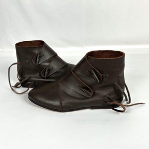 May include: A pair of brown leather ankle boots. The boots have a rounded toe, a folded-over cuff, and are secured with leather ties and straps. The boots appear to be handmade and are suitable for historical reenactment or costume.