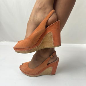 May include: A pair of orange suede slingback wedge shoes featuring an open-toe design and a cork-textured wedge heel. The shoes include a thin adjustable ankle strap with a small metal buckle and visible white stitching along the cork panel.