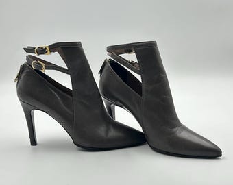 Puede incluir: Un par de botines de cuero marrón oscuro con punta y tacones de aguja. Los botines presentan un diseño recortado en los laterales y correas en el tobillo con hebillas doradas. Los tacones miden aproximadamente 10 cm.