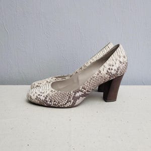 Pode incluir: Um par de sapatos de salto alto com padrão de pele de cobra em tons de marrom e branco. Os saltos são de bloco e são feitos de madeira.
