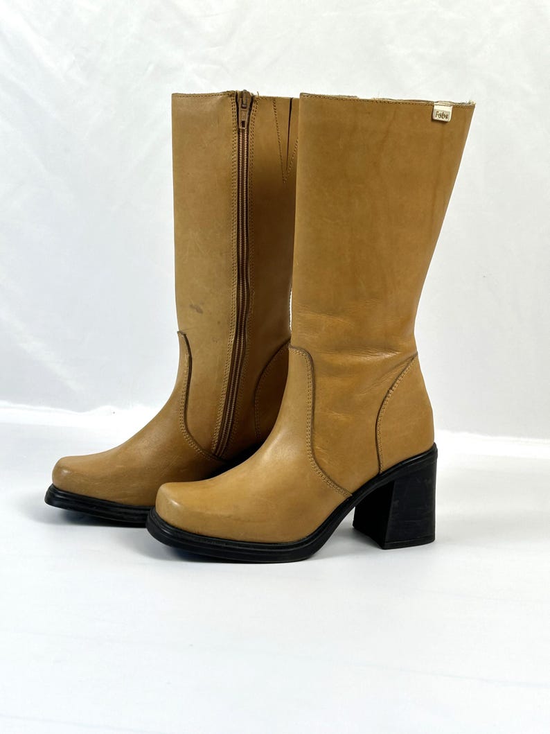 Puede incluir: Un par de botas de cuero color canela con punta cuadrada y tac&oacute;n de bloque negro. Una bota tiene una cremallera lateral. Las botas tienen suela de plataforma, sobre fondo blanco.