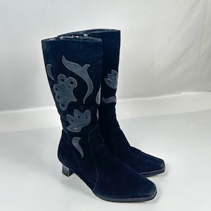 Puede incluir: Un par de botas de gamuza negras con diseños florales recortados. Las botas tienen una punta puntiaguda y un tacón bajo.